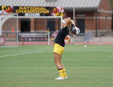 A soccer player in yellow shorts and black shirt prepares to throw a soccer ball during a game. The background includes a banner that reads 'National Champions 2005' and a brick building.