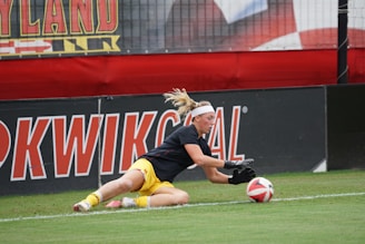 A soccer player in action on the field is diving to catch a ball. The athlete is wearing a black jersey and yellow shorts, with gloves on their hands. The background shows large banners with text and a part of a goal post.