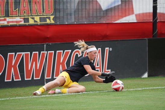 A soccer player in action on the field is diving to catch a ball. The athlete is wearing a black jersey and yellow shorts, with gloves on their hands. The background shows large banners with text and a part of a goal post.