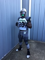 A football player in full gear stands in front of a corrugated metal wall. The uniform is primarily gray and white, and the player is wearing a helmet and gloves. The number 28 is prominently displayed on the jersey.