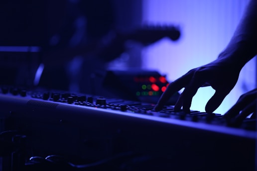 A close-up of a person's hand playing a keyboard instrument in a dimly lit setting, with a focus on the fingers pressing the keys. The background is blurred, showing blue and purple hues with some glowing lights.