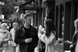 A street scene featuring a group of people walking along a sidewalk. In the foreground, a man and a woman are engaged in conversation, both holding cups, suggesting a sense of casual interaction. The background shows a mix of pedestrians and store signs, adding to the urban atmosphere.