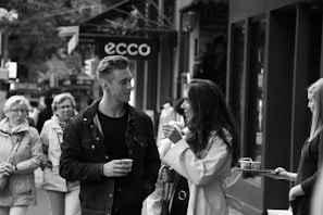A street scene featuring a group of people walking along a sidewalk. In the foreground, a man and a woman are engaged in conversation, both holding cups, suggesting a sense of casual interaction. The background shows a mix of pedestrians and store signs, adding to the urban atmosphere.