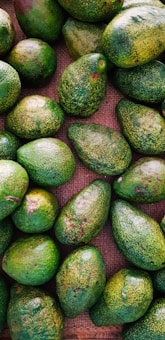 A collection of ripe avocados lies on a textured burlap surface. The avocados are predominantly green with some brownish spots, varying slightly in shape and size. They are densely arranged, covering most of the image in a natural, organic pattern.
