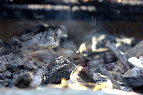 Charred pieces of coal and ash lie scattered, with a few embers glowing faintly under a soft haze of smoke. The texture of the charcoal is rough and burnt, with some pieces showing lighter gray ash and others darker black surfaces.