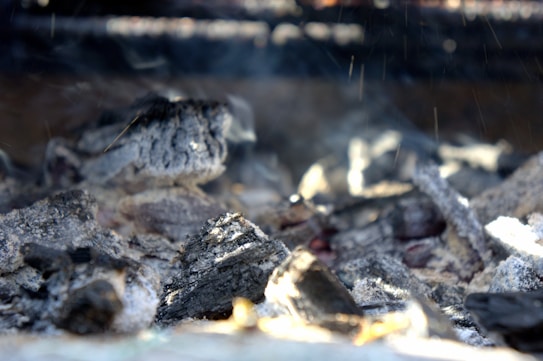 Charred pieces of coal and ash lie scattered, with a few embers glowing faintly under a soft haze of smoke. The texture of the charcoal is rough and burnt, with some pieces showing lighter gray ash and others darker black surfaces.
