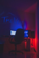 A dimly lit room with a dark ambiance features a computer setup. A monitor and a glowing CPU are placed on a desk, with a swivel chair positioned in front. The words 'The Void' are illuminated on the wall in vibrant blue and red neon lights, creating a moody atmosphere.