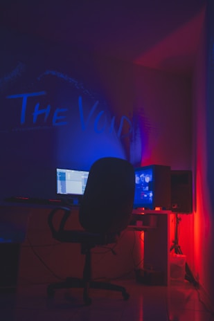 A dimly lit room with a dark ambiance features a computer setup. A monitor and a glowing CPU are placed on a desk, with a swivel chair positioned in front. The words 'The Void' are illuminated on the wall in vibrant blue and red neon lights, creating a moody atmosphere.