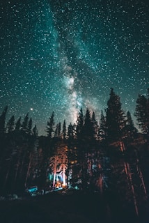 A breathtaking view of the starry night sky with the Milky Way clearly visible, towering over a forest of tall pine trees. A small campsite with a glowing tent and a car can be seen nestled among the trees, illuminated by soft lighting.
