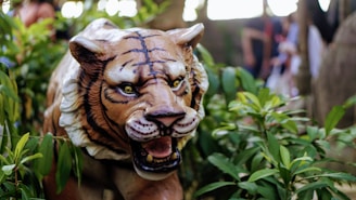 A detailed depiction of a realistic tiger statue amidst lush green foliage. The tiger has a fierce expression with its mouth open, revealing sharp teeth. Its eyes are wide and appear menacing, adding to the lifelike appearance of the statue. The surrounding environment includes dense greenery, creating a jungle-like atmosphere.