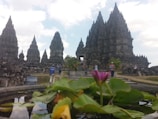 A historic temple complex with pointed, intricate stone structures stretches towards the sky, surrounded by lush greenery and a pond in the foreground. People are casually walking around, indicating a tourist attraction or archaeological site.