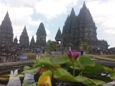 A historic temple complex with pointed, intricate stone structures stretches towards the sky, surrounded by lush greenery and a pond in the foreground. People are casually walking around, indicating a tourist attraction or archaeological site.
