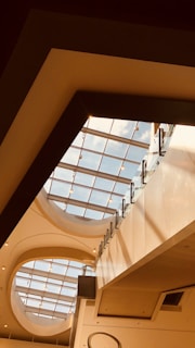 A modern architectural interior with a focus on a large skylight. The ceiling features geometric patterns and clean lines, with natural light streaming through the glass panes. The materials used include glass, metal, and plaster.