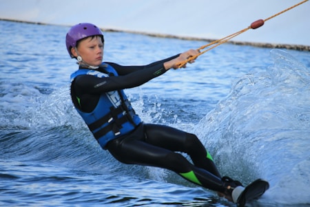 A young person is engaged in water skiing or wakeboarding on a body of water while wearing a life jacket, a wetsuit, and a purple helmet. The individual is holding onto a yellow ski rope handle, and is captured leaning back slightly as the water splashes around them.