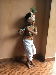 A child dressed in traditional Indian attire is holding a flute. The outfit includes a decorative headpiece with peacock feathers and brightly colored embellishments, along with a white dhoti. The setting appears to be indoors with a plain wall and tiled floor.