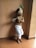 A child dressed in traditional Indian attire is holding a flute. The outfit includes a decorative headpiece with peacock feathers and brightly colored embellishments, along with a white dhoti. The setting appears to be indoors with a plain wall and tiled floor.