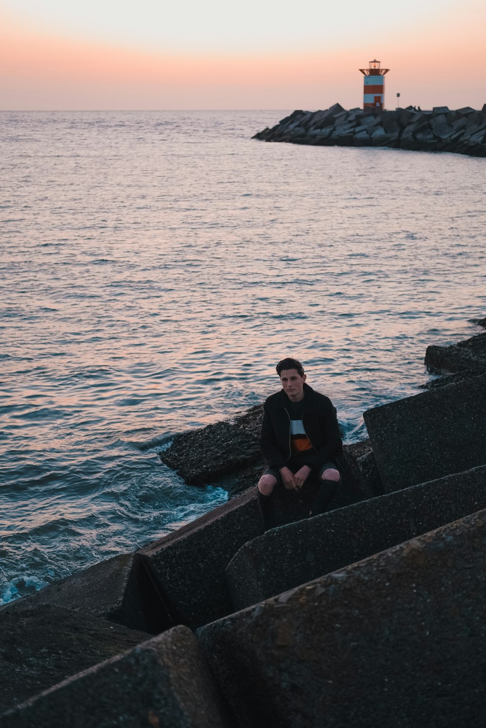 man sitting on concrete pavement beside body of water with lighthouse ahead