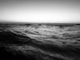 A black and white photograph captures the dynamic motion of ocean waves under a subtle gradient sky. The waves appear to be in constant movement, creating a sense of energy and fluidity across the scene.