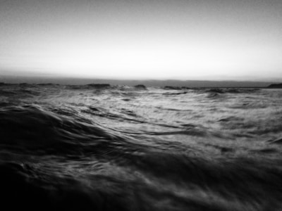 A black and white photograph captures the dynamic motion of ocean waves under a subtle gradient sky. The waves appear to be in constant movement, creating a sense of energy and fluidity across the scene.