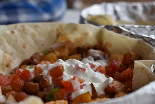 A close-up of a loaded tortilla filled with diced tomatoes, beans, potatoes, and drizzled with sour cream, sitting on a sheet of aluminum foil.