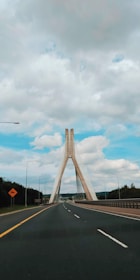A modern suspension bridge spans across a wide view, with its distinct, tall triangular pillars rising into a cloudy sky. The bridge is part of a road with multiple lanes, bordered by green landscapes and tall street lamps. A caution sign is visible on the left side of the road.