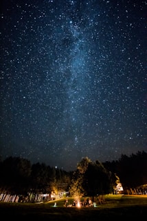 A night sky filled with countless stars is seen above a serene landscape. In the foreground, there is a small campfire surrounded by a group of people, partially obscured by tall trees. The distant lights of a house or cabins are visible through the trees, creating a warm contrast to the cool tones of the starlit sky.