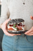 A person holding a unique hamburger with a black sesame seed bun. The burger includes visible ingredients such as sliced tomatoes, onion rings, fresh greens, cheese, and a patty. The individual is wearing a light-colored top and denim jeans.