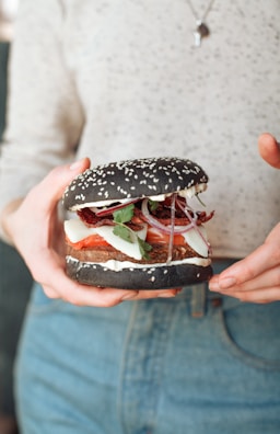 A person holding a unique hamburger with a black sesame seed bun. The burger includes visible ingredients such as sliced tomatoes, onion rings, fresh greens, cheese, and a patty. The individual is wearing a light-colored top and denim jeans.