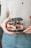 A person holding a unique hamburger with a black sesame seed bun. The burger includes visible ingredients such as sliced tomatoes, onion rings, fresh greens, cheese, and a patty. The individual is wearing a light-colored top and denim jeans.