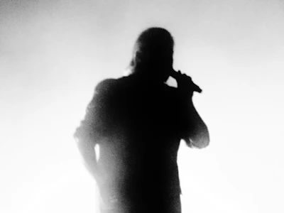 Shadowy silhouette of a person speaking into a microphone, surrounded by deep black and gray tones.