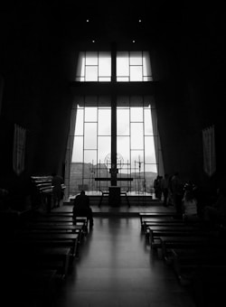 A dimly lit, monochromatic interior of a church with a large cross silhouetted against a tall window. There are benches lined up leading towards the altar, and a few people are seated or standing near the front.