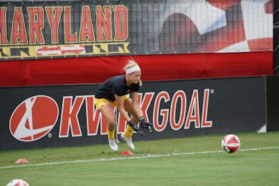 A soccer player wearing a black shirt and yellow shorts practices on a field, ready to catch a soccer ball. The field is bordered by advertising boards, one with the word 'KWIKGOAL.' The player is wearing gloves and positioned between red cones, with a banner that reads 'MARYLAND' in the background.