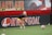 A soccer player wearing a black shirt and yellow shorts practices on a field, ready to catch a soccer ball. The field is bordered by advertising boards, one with the word 'KWIKGOAL.' The player is wearing gloves and positioned between red cones, with a banner that reads 'MARYLAND' in the background.