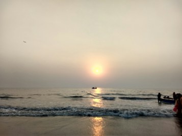 The image portrays a serene seascape during sunset, with a glowing sun hovering just above the horizon, casting a golden reflection on the calm waters. A small boat is visible in the water near the horizon, while waves gently roll onto the shore. A few people are gathered on the beach, engaging in leisure activities. The sky has a gradient of soft hues, giving a peaceful ambiance to the scene.