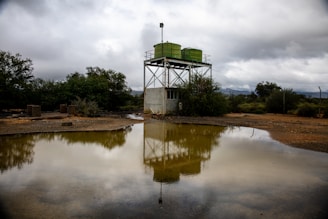 A metal water tank structure with two large green tanks is reflected in a muddy pool of water. The scene is set in an open, arid landscape with overcast skies and dense bushes surrounding the area.