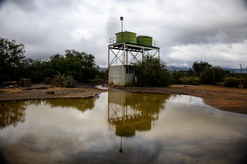 A metal water tank structure with two large green tanks is reflected in a muddy pool of water. The scene is set in an open, arid landscape with overcast skies and dense bushes surrounding the area.