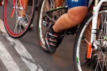 Cyclists are in close proximity, with focus on the lower body and bicycles. The image captures a busy and energetic scene likely from a race or group ride. The emphasis is on the cycling shoes, pedals, and the bicycles' frames and wheels.