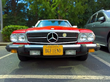 A classic red Mercedes-Benz car with a Virginia license plate reading 'BGF 1END' parked in a parking lot. The car features a chrome grille, round headlights, and an antique plate, indicating its vintage status. It is surrounded by greenery in the background and a modern car to the right.