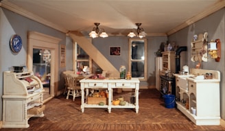 A detailed miniature kitchen scene featuring a rustic interior with white wooden furniture. The room includes a central table holding various items like vegetables and jars, a stove with pans, and shelves with decorative plates and jars. A wicker chair and a desk are present near the doorway. The floor is a patterned wood, and there are warm ceiling lights.