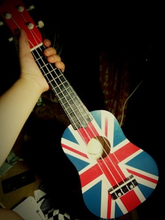 A hand holding a ukulele decorated with the Union Jack, the flag of the United Kingdom. The background is dark, focusing attention on the instrument. The ukulele's body features the red, white, and blue colors of the flag, with four strings running along the fretboard.