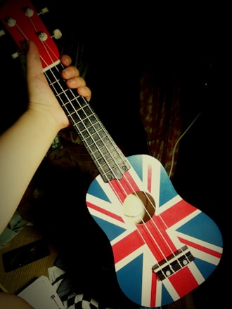 A hand holding a ukulele decorated with the Union Jack, the flag of the United Kingdom. The background is dark, focusing attention on the instrument. The ukulele's body features the red, white, and blue colors of the flag, with four strings running along the fretboard.