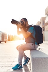 A person with a large camera lens is sitting on a stone ledge, focusing on taking a photograph. They are wearing casual athletic clothing and a full backpack, with the sun setting in the background casting a warm glow.