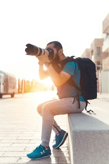 A person with a large camera lens is sitting on a stone ledge, focusing on taking a photograph. They are wearing casual athletic clothing and a full backpack, with the sun setting in the background casting a warm glow.