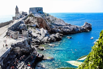 A scenic coastal landscape featuring rocky cliffs with an ancient fortress and a church overlooking the vibrant blue sea. People are seen walking along the stone pathways, while others swim in the clear waters. Lush green plants are visible in the foreground, contrasting with the rugged stone structures and the calm ocean.