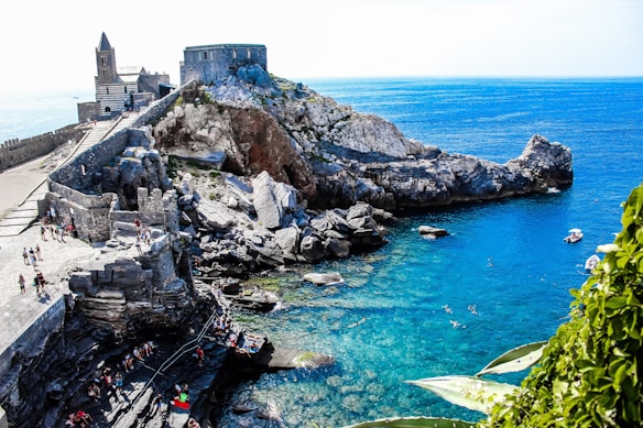 A scenic coastal landscape featuring rocky cliffs with an ancient fortress and a church overlooking the vibrant blue sea. People are seen walking along the stone pathways, while others swim in the clear waters. Lush green plants are visible in the foreground, contrasting with the rugged stone structures and the calm ocean.