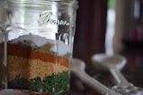 A glass jar displays multiple layers of spices, including green herbs, coarse orange and brown powders, and white granules. The container sits on a kitchen counter next to several measuring spoons.