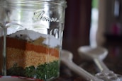 A glass jar displays multiple layers of spices, including green herbs, coarse orange and brown powders, and white granules. The container sits on a kitchen counter next to several measuring spoons.