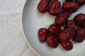 A group of reddish-brown dates are arranged on a round, light-toned ceramic plate, placed over a white fabric background. The dates have a wrinkled texture and are positioned on the right side of the plate.