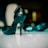 A glossy black high-heeled shoe is prominently displayed, standing upright on a shiny surface. In the blurred background, another similar shoe is visible. The image has a soft focus effect that emphasizes the shoe in the foreground.