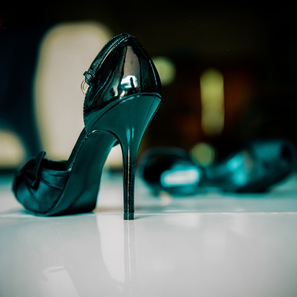 A glossy black high-heeled shoe is prominently displayed, standing upright on a shiny surface. In the blurred background, another similar shoe is visible. The image has a soft focus effect that emphasizes the shoe in the foreground.
